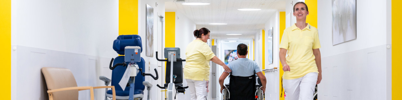 Schlaganfall-Patient im Rollstuhl wird begleitet auf Station von der Pflegemitarbeiterin